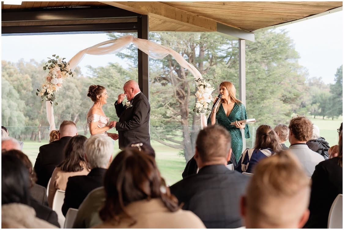 Katie speaking into a microphone at the ceremony arch while the groom wipes away a tear and guests look on